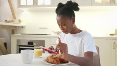 Ellerinde akıllı telefonla kahvaltı, ev mutfağında cep telefonuyla internette sörf yapan genç siyahi kadın, alet bağımlısı birinin portresi, Afrikalı Amerikalı etnik kökenler.