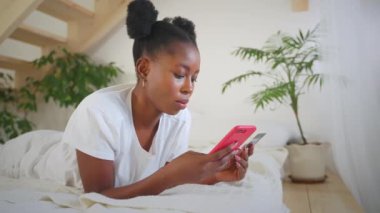 Çevrimiçi ödeme yapan Afrikalı Amerikalı bir kız. Banka kartıyla ödeme yapıyor. Evde yatakta uzanan akıllı telefonlara numara ekliyor. At kuyruklu kadın alıcı telefon ekranına bakıyor. İnternet pazarında internet alışverişi.