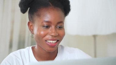 Gülümseyen Afro-Afrikalı bir kadın dizüstü bilgisayara bakıyor çevrimiçi sohbet ediyor, görüntülü sohbet yapıyor. Kıvırcık at kuyruklu serbest çalışan kız ev ofisinden bilgisayar başında konuşuyor..