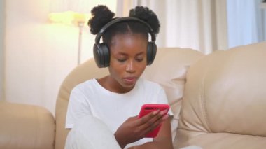 Rahat kız kulaklıkla müzik dinliyor. Afro-amerikan kadın müzik hayranı favori melodilerin tadını çıkarıyor evde oturmuş akıllı telefon ekranına bakıyor. Eğlenmek için telefon kullanan bir kadın.