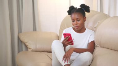 Telefonla bilgi okuyan bir kadın. Kıvırcık at kuyruklu Afrikalı Amerikalı kız sosyal medya sayfalarında akıllı telefondan bilgi arıyor. Evdeki koltukta oturuyor. İnternette yaşamak, internet bağımlılığı.