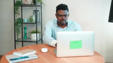 Ciddi bir Afro-Amerikalı, gözlüklü, bilgisayarlı ve merkez ofisteki masada oturuyor. Serbest iş, dijital göçebeler konsepti. İçerik oluşturan odaklanmış adam blogcusu yazı yazıyor.