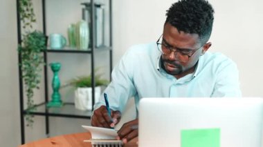 Serbest çalışan, bilgisayarla çalışan, merkez ofiste portre çizen, not defterine not alan Afrikalı Amerikalı erkek ve dizüstü bilgisayara bakan, uzak çalışma modu için iş yeri, dijital göçebe.