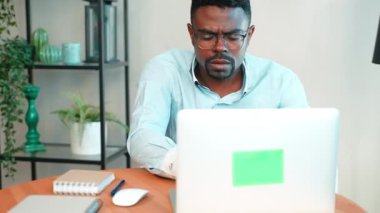Ciddi Afrika kökenli Amerikalı serbest çalışan bir adam not defterine önemli bilgiler yazıp ev ofisinin bilgisayarındaki bilgisayar ekranına bakıyor. Dijital göçebe, uzak görev konsepti.