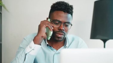 Ciddi bir Afro-Amerikan iş adamı laptopla çalışırken dahili ofisteki akıllı telefonu arıyor. Portre odaklı adamın iş yerinde sorunları var. Uzak bağımsız iş, dijital göçebe konsepti.