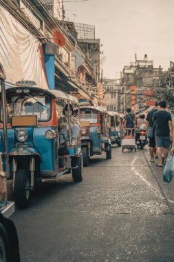 Busy urban street in Southeast Asia featuring traditional tuk-tuks lined up in traffic. The scene captures the vibrant local culture, with bustling markets, pedestrians, and vintage architecture. clipart