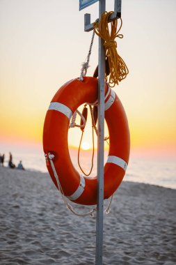 A vibrant sunset illuminates the beach, framing a bright orange lifebuoy against the tranquil waves. clipart