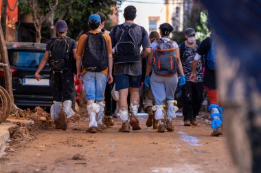 Fotoğraf Alfafar, Valencia, İspanya 'daki selin sonuçlarını gösteriyor. Bir grup gönüllü, selden etkilenmiş bir bölgede, sırt çantaları ve koruyucu ekipmanlarla çamurlu bir sokakta yürüyor..
