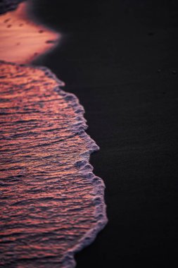 The gentle waves leave a trail of foam on the darkened sand, reflecting the vibrant hues of pink and orange from the setting sun. clipart