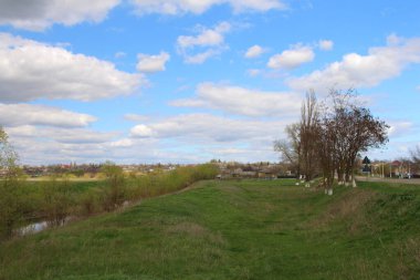 Nehirli bir manzara, ağaçlar, çimenler, ve bulutlu güzel bir gökyüzü.
