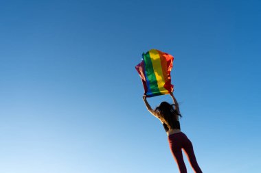 LGBT bayrağını rüzgarda tutan kadının arka planında mavi bir gökyüzü var.