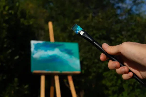 Brush seen from up close with an easel with a canvas of contemporary art in nature in the background