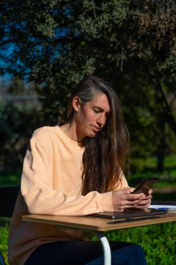 Orta yaşlı, beyaz bir kadın evinin bahçesinde masa ve cep telefonuyla çalışıyor.