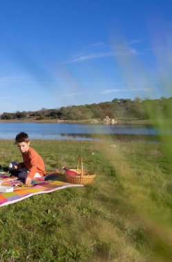 Çocuk göl kenarında piknik yapıyor, piknik sepeti ve turuncu tişörtle.