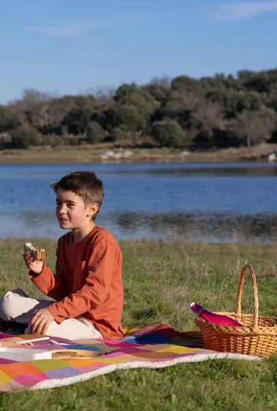 Çocuk turuncu tişörtle gölde piknik yaparken sandviç yiyor.
