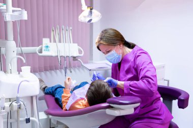 Dişçi koltuğunda yatan bir çocuğun dişlerine bakan dişçi kadın.