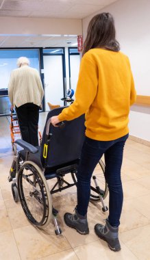 Kıdemli kadın tekerlekli sandalye taşıyan bir tekerlekli sandalyeyle hastanede yürüyor.
