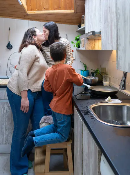 stock image Couple of two lesbian women kissing while cooking with their son in the kitchen of their house. LGBTTTQIA rights