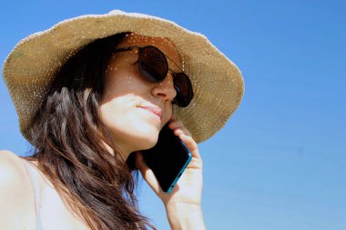 Cep telefonunda güneş gözlüklü ve şapkalı bir kadının yüzü konuşuyor.