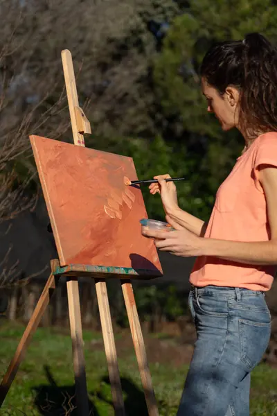 Artist woman painting with brush a canvas on an easel in nature with peach colors