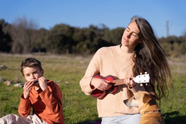 Anne ve oğul doğada enstrüman, ukulele ve mızıka çalıyorlar.