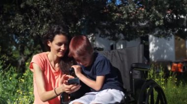 Anne ve oğlu tekerlekli sandalyede doğada cep telefonuna bakıyorlar.