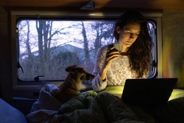 Woman in pajamas using the computer in the bed of a motorhome with her dog clipart