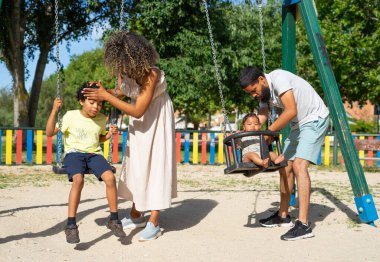 İki çocuklu Latin bir aile, 8 yaşında bir oğlan ve 6 aylık bir bebek çocuk parkında salıncakta sallanıyor.