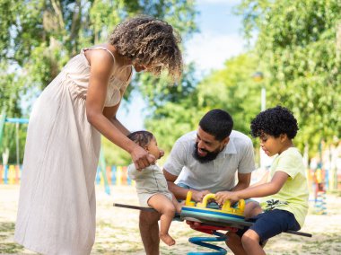 Latin bir aile, çocukları, bir oğlan ve bir bebekle oyun parkında birlikte oynuyor.
