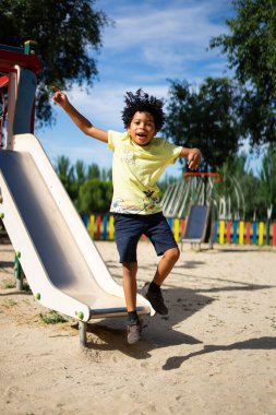 7 yaşında, Latin bir çocuk, afro saçlı, oyun parkında kayaktan atlıyor.