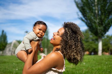 6 aylık Latin bir bebek, annesi tarafından bir parkta saçlarıyla tutulurken kameraya bakıyor.