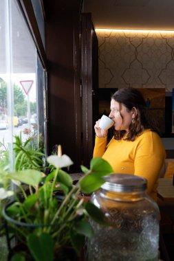 Orta yaşlı, beyaz bir kadın bir kafede tek başına kahve içerken pencereden dışarı bakıyor.