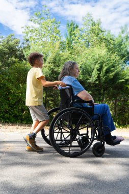 Çocuk büyükannesini tekerlekli sandalyeyle yürüyüşe çıkarıyor.
