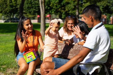 Bebekli Latin bir aile parktaki bankta kafiyeli şarkı söylüyor.