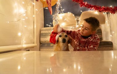 Noel kıyafetleri ve Noel süslemeleri ile bir karavanda köpeği olan bir çocuk. Çirkin