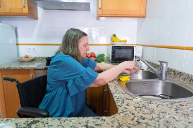 60 yaşından büyük bir kadın tekerlekli sandalyede evinin mutfağında bulaşık yıkıyor.