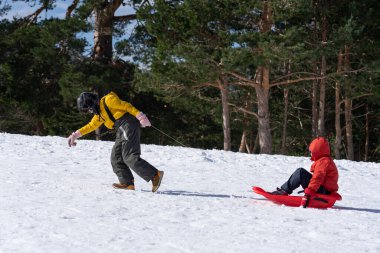 Two children playing in the snow with a sled, one pulling the other clipart