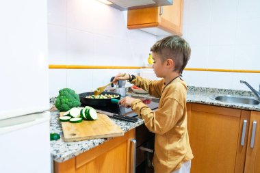 Beyaz çocuk evdeki mutfakta tavada sebze pişirmeyi öğreniyor.