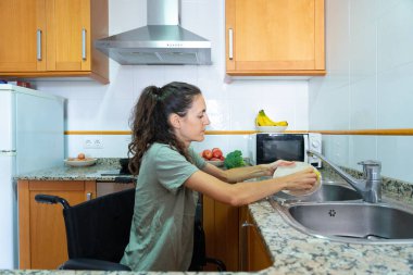 Tekerlekli sandalyedeki beyaz kadın evde bulaşık yıkıyor.