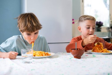 Children eating spaghetti in the kitchen at home clipart