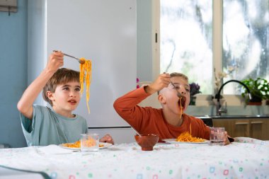 İki genç kardeş domates soslu spagetti yerken evlerinde yemek yerken neşeli ve neşeli ifadeler sergiliyorlar.