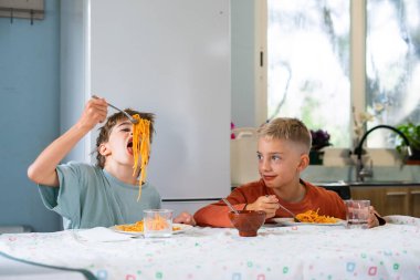 Two brothers enjoying a spaghetti meal, one playfully slurping a long strand while the other smiles, showcasing childhood joy during mealtime clipart