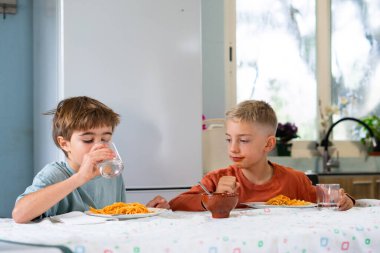 İki genç kardeş domates soslu spagetti yemeğinin tadını çıkarıyorlar. Susuzluklarını açık mutfak ortamında bir bardak suyla gideriyorlar.