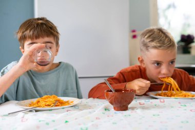 İki beyaz çocuk evde spagetti yiyor, biri içme suyu, diğeri ise yemek yemeye odaklanmış.