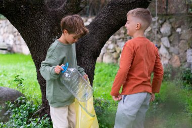 İki genç çocuk, çevre bilincini artırmak için sarı bir çöp torbasına plastik bir şişe koyuyorlar.