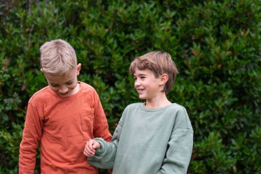 İki kardeş yeşil bir çitin önünde gülümsüyor.