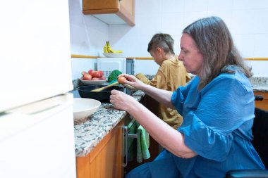 Senior woman in wheelchair preparing food with her grandson helping her in the kitchen clipart