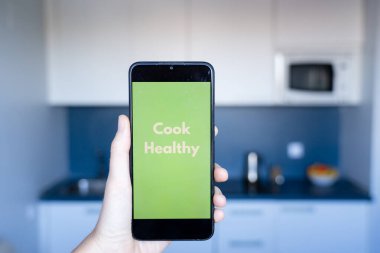 Elinde akıllı bir telefon olan biri, yeşil ekran kullanıyor ve modern bir mutfakta sağlıklı yemek pişiriyor, teknolojiyle sağlıklı bir yaşam tarzını destekliyor.