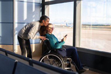 Havaalanında akıllı telefon kullanan tekerlekli sandalyedeki bir yolcuya yardım eden bir kadın.