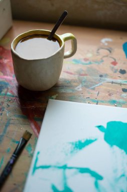 Coffee mug sits on a colorful, paint-splattered table next to a canvas with abstract teal brushstrokes, suggesting a pause in an artist's work clipart